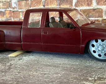 Peut inclure: Un camion rouge bordeaux avec un intérieur blanc et de grandes roues argentées. Le camion est garé sur une surface en béton gris.