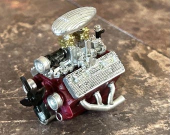 May include: A miniature model of a classic car engine. The engine features a maroon block, silver and gold components, and a silver air cleaner. The model is highly detailed, showcasing the intricate design of the engine.