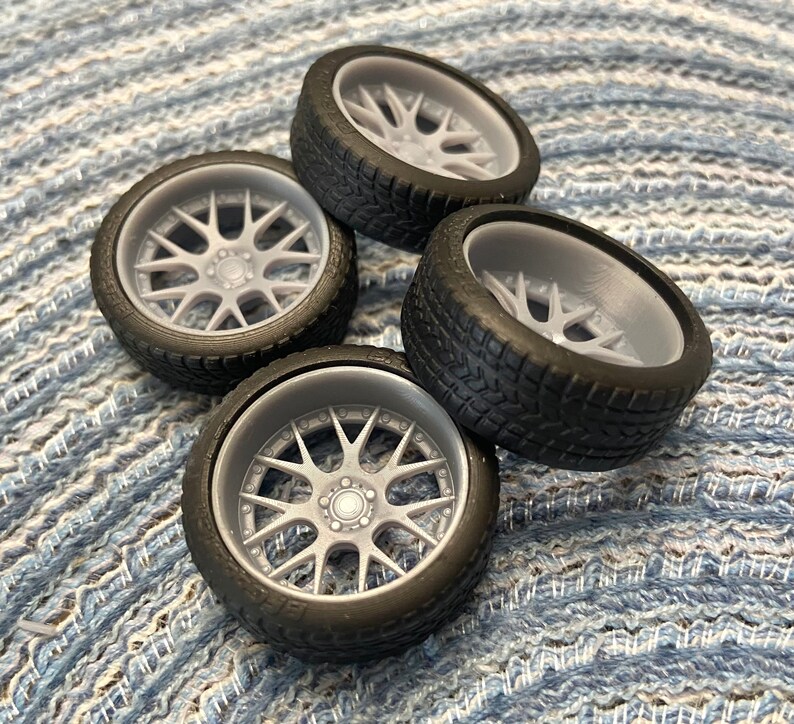 Puede incluir: Cuatro neum&aacute;ticos de caucho negro con llantas grises de radios detallados. Los neum&aacute;ticos est&aacute;n dispuestos en una pila sobre una superficie con un patr&oacute;n azul y blanco.