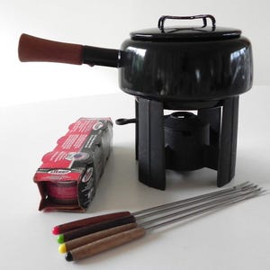 May include: Black fondue pot with a wooden handle and a black metal stand. The pot has a lid and a small metal bowl underneath. There are six fondue forks with wooden handles and a box of fondue fuel.