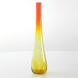 Pode incluir: Um vaso de vidro alto e fino com um efeito degradê. O topo é de um laranja vibrante que se funde em um amarelo brilhante em direção à base. O vaso tem um pescoço longo e estreito e uma base ligeiramente arredondada.