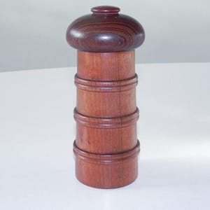 Może przedstawiać: Drewniany młynek do pieprzu z ciemnobrązową górą i cylindrycznym korpusem. Korpus składa się z ułożonych w stos sekcji drewna, każda z ozdobnym rowkiem. Młynek ma ciepły, czerwonobrązowy kolor.