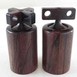 May include: Two wooden salt and pepper shakers. The shakers are dark brown with a visible wood grain. One shaker has a cross-shaped top, and the other has a rectangular top with two holes. The shakers are cylindrical in shape.