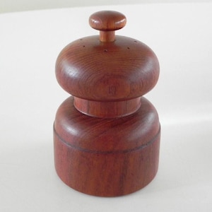 Może przedstawiać: Drewniany młynek do pieprzu o bogatym, czerwonobrązowym wykończeniu. Młynek ma zaokrąglony korpus, środkową część i górę z małym pokrętłem. Klasyczny design do użytku w kuchni.