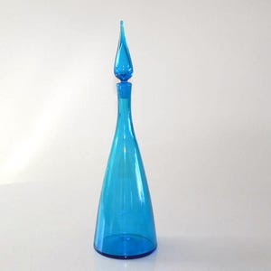 Op de afbeelding: Een hoge, turquoise glazen karaf met een druppelvormige stop. De fles heeft een conische vorm en een lange, slanke hals. De stop heeft een bijpassende turquoise kleur en is ook van glas gemaakt.