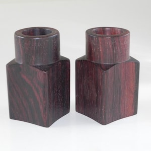 Może przedstawiać: Dwa ciemne drewniane świeczniki z kwadratową podstawą i okrągłym wierzchołkiem. Świeczniki są wykonane z ciemnego drewna o czerwonawej barwie.