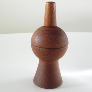 Może przedstawiać: Drewniana solniczka lub pieprzniczka z pękatym korpusem i stożkową górą. Shaker wykonany jest z ciepłego, czerwonobrązowego drewna i ma gładkie, wypolerowane wykończenie. Na górze znajduje się szereg małych otworów do dozowania zawartości. Ma około 15 cm wysokości.