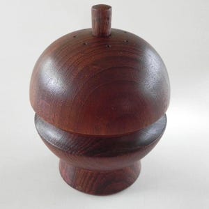 Może przedstawiać: Ciemnobrązowy, drewniany młynek do pieprzu z zaokrągloną górą i małym, cylindrycznym uchwytem. Młynek ma pękatą podstawę i gładkie, wypolerowane wykończenie.