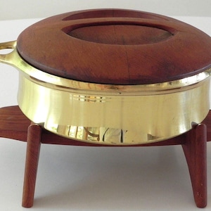 May include: A brass serving dish with a wooden lid and stand. The dish is round and has a gold finish. The lid is made of dark wood and has a handle. The stand is made of wood and has three legs.