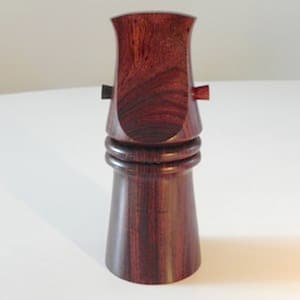 Może przedstawiać: Korek do butelki z ciemnego drewna o unikalnym wzorze. Korek ma zaokrąglony wierzchołek i podstawę z pierścieniem wokół.