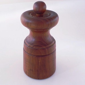 Może przedstawiać: Drewniany młynek do pieprzu w ciemnobrązowym wykończeniu. Młynek ma cylindryczną podstawę, pękatą środkową część i zaokrągloną górę z małym pokrętłem. Widoczne usłojenie drewna. Przyrząd kuchenny.