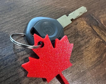 May include: A red maple leaf keychain with a silver key ring and a car key. The keychain is made of a textured material and is a vibrant red color. The key is silver and black.