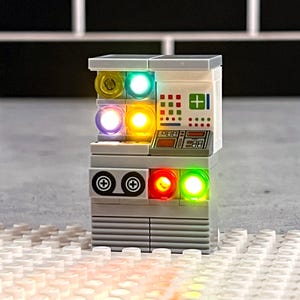 May include: A white Lego model of a computer with a cassette tape player and a control panel with lights. The lights are red, green, yellow, and blue.