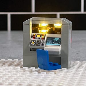 May include: A gray Lego model of a computer workstation with a blue chair, a computer screen with a radar display, and a control panel with buttons and lights. The lights are on and glowing yellow.