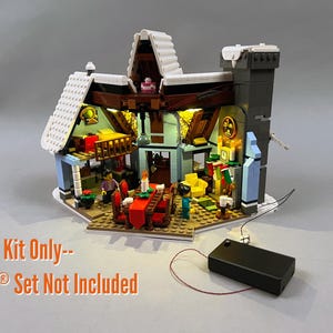 Puede incluir: Un set de construcción en miniatura con techo nevado, iluminación interior, mesa de comedor con camino de mesa rojo, sillón amarillo y chimenea con calcetines. Una caja de baterías negra está conectada a la estructura mediante cables finos.