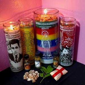 May include: An assortment of lit glass candles with various labels and designs. One candle features a portrait and the text "Niño Fidencio Constantino." Another displays a cross and the words "Good Luck." Other candles have the text "La Santisima Muerte" and "Hummingbird Nectar of Love."