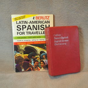 May include: Two paperback books, one red and one with a yellow cover. The yellow book is titled "Latin-American Spanish for Travel" with the Berlitz logo. The red book is titled "Collins Deutsch-Englisch English-German Dictionary".