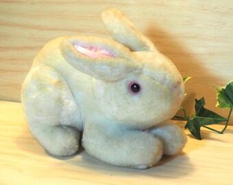 May include: A plush beige bunny rabbit with pink ears and black eyes. The bunny is sitting on a wooden surface.