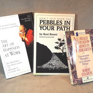 May include: Three hardcover books with colorful covers. The book on the left is titled "The Art of Happiness at Work" by His Holiness the Dalai Lama and Howard C. Cutler, M.D. The middle book is titled "Don't Trip Over the Pebbles in Your Path" by Roni Bissett. The book on the right is titled "All I Really Need to Know I Learned in Kindergarten" by Robert Fulghum.