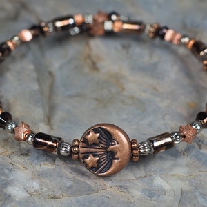 May include: A copper bracelet with a moon and stars charm. The bracelet is made with brown, black, and silver beads.