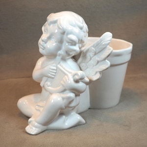 May include: A white ceramic cherub figurine playing a harp, with detailed wings and curly hair. The cherub is seated next to a small, matching white planter. The figurine has a glossy finish and is set against a neutral background.