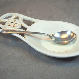 May include: A white ceramic spoon rest with a decorative handle and a silver spoon. The spoon rest has a distressed finish with brown accents. The spoon is resting in the center of the rest.