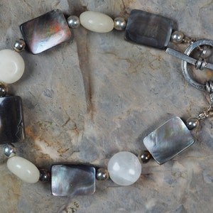 Könnte beinhalten: Ein Armband mit rechteckigen, irisierenden grauen und schwarzen Steinen, die mit weißen ovalen und runden Perlen abwechseln. Silberfarbene Metallperlen und ein Knebelverschluss vervollständigen das Design. Das Armband ist etwa 19 cm lang.