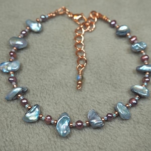 May include: A delicate bracelet featuring iridescent blue-gray keshi pearls and small, dark pink beads. The bracelet is strung on a copper-colored chain with an adjustable clasp. A beautiful piece of jewelry.