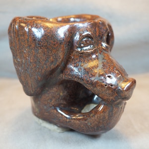 May include: A brown ceramic dog head planter with a speckled glaze. The planter has a wide, open mouth and a detailed face.