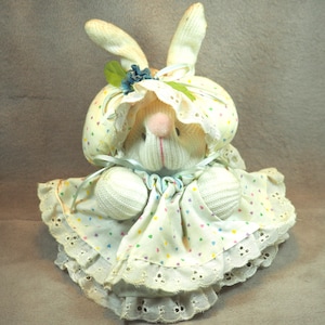 Puede incluir: Un conejito de calcetín blanco con un vestido blanco con lunares de colores y un borde de encaje blanco. El conejito tiene un gorro de encaje blanco con una hoja verde y una flor azul.