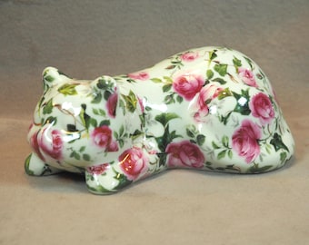 May include: A ceramic cat figurine in a reclining pose, adorned with a floral pattern of pink roses and green leaves on a cream background. The cat's ears and paws are visible, showcasing the detailed design.