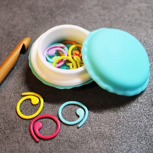 May include: A turquoise blue plastic container shaped like a macaron with a white lid. The container is open and filled with colorful plastic stitch markers. Three stitch markers are on the gray surface next to the container.