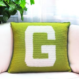 May include: A green crocheted cushion with a white letter "G" in the centre.