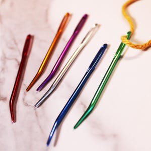 May include: A set of seven colorful needles with a curved tip. The needles are made of metal and have a shiny finish. The needles are arranged in a row on a white surface.
