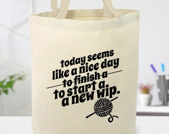 May include: A white canvas tote bag with black text that reads "today seems like a nice day to finish a to start a. a new wip."  The text is crossed out and a yarn ball and knitting needle are pictured below the text.
