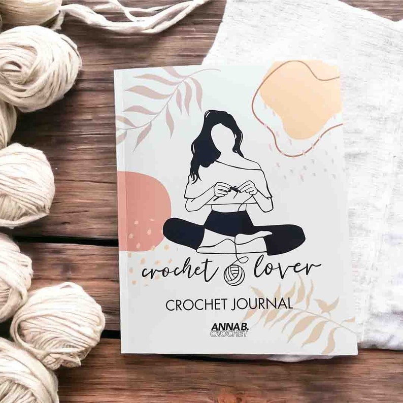 Pu&ograve; includere: Un diario di uncinetto bianco con un'illustrazione in bianco e nero di una persona che fa l'uncinetto. Il diario ha il testo "crochet lover" e "CROCHET JOURNAL" sulla copertina. Il diario ha anche il testo "ANNAB. CROCHET" sulla copertina.