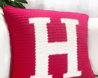 Puede incluir: Un cojín decorativo de ganchillo en color magenta vibrante. El cojín presenta una gran letra "H" blanca en el centro. El cojín es de forma cuadrada y parece estar hecho de hilo suave.