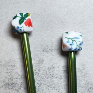 May include: Two knitting needle stoppers. Each stopper has a white, cube-shaped top with a floral pattern in red, blue, and green. The needles are a metallic green color. The stoppers are designed to prevent stitches from falling off the needles.