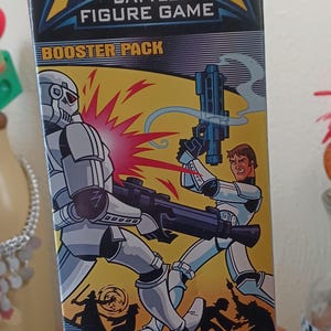 May include: An AttakTix Star Wars Battle Figure Game booster pack box featuring a graphic illustration of a white armored trooper and a human character in combat. The packaging includes the text AttakTix, Booster Pack, 3 Figures Included, and Star Wars.