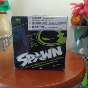 May include: A black and green cardboard box for McFarlane Toys Spawn Series 1 mini trading figures. The front features the Spawn logo, a skull icon, and safety warnings in English, French, and Spanish, noting it is for ages 5 and up.