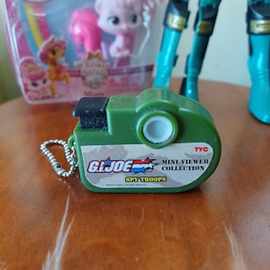 Può includere: Un portachiavi G.I. Joe Mini Viewer Collection Spy Troops verde oliva con un pulsante superiore nero, una lente circolare bianca e una catenella a sfere argentata. L'etichetta riporta il logo G.I. Joe, il testo TYC e l'anno 2003.