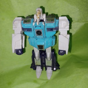 May include: A vintage action figure in a robot design. The toy features a light blue torso, white arms, and dark blue and black legs. The head is a light tan color. The toy is standing on a green surface.