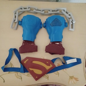 May include: A set of blue and maroon plastic wrist gauntlets with embossed Superman logos, a grey plastic chain link accessory, and a red and gold Superman chest emblem attached to a blue elastic strap for a costume accessory set.