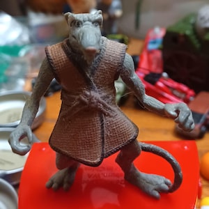May include: A gray anthropomorphic rat figurine wearing a brown textured vest with a tied waist belt. The character has pointed ears, a long tail, and clawed hands. The figure stands in an upright position with a neutral facial expression.