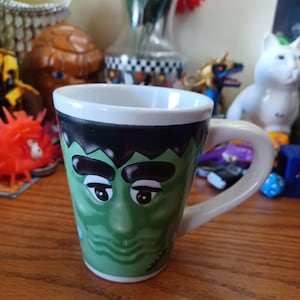 May include: A ceramic mug featuring a green, stylized face with black hair, thick eyebrows, and a small scar detail on the side. The mug has a white interior, a white handle, and a tapered shape, showcasing a character-inspired design.
