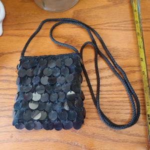 May include: A black crossbody bag covered in circular black sequins with a braided black cord strap. A yellow measuring tape is positioned to the right, showing the bag is approximately 7 inches tall and 6 inches wide at the base.