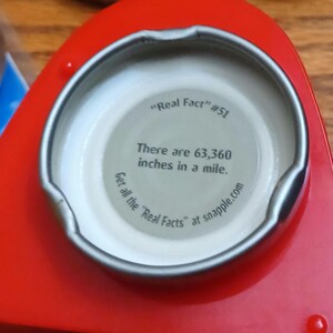 May include: A metal bottle cap with a red exterior and a white interior. The inside features the text "Real Fact" #51, stating "There are 63,360 inches in a mile." and "Get all the "Real Facts" at snapple.com" printed in black ink.