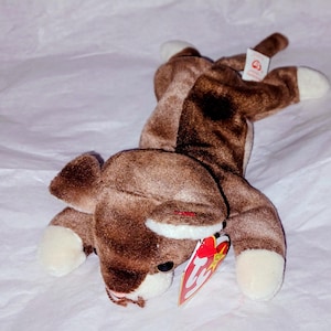 Peut inclure: Un jouet en peluche brun en forme de renne avec des accents blancs sur les sabots et les bois. Le jouet a une étiquette rouge attachée à l'oreille. Le renne est allongé sur une surface blanche.