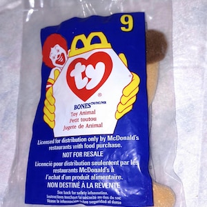 May include: Sealed blue and white Ty Animal toy in a plastic bag. The bag features the McDonald's logo, the number 9, and text in English, French, and Spanish. The toy is tan and brown.