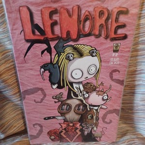 May include: A comic book titled "Lenore" with a pink background and a cartoon illustration of a girl with blonde hair and skull accents. The cover features various other cartoon characters and the text "#8".